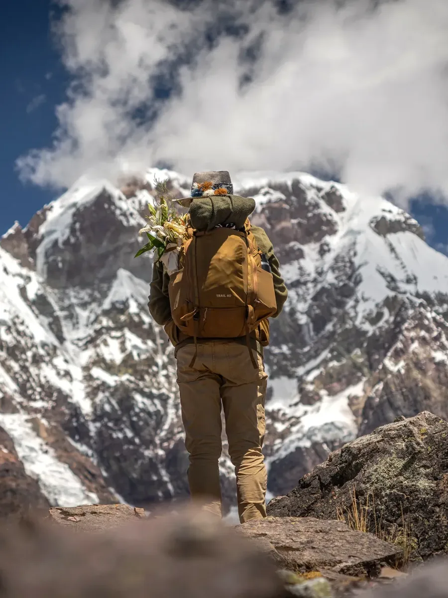 A pilgrim with a loaded pack and a bundle of flowers walks toward a glaciated peak