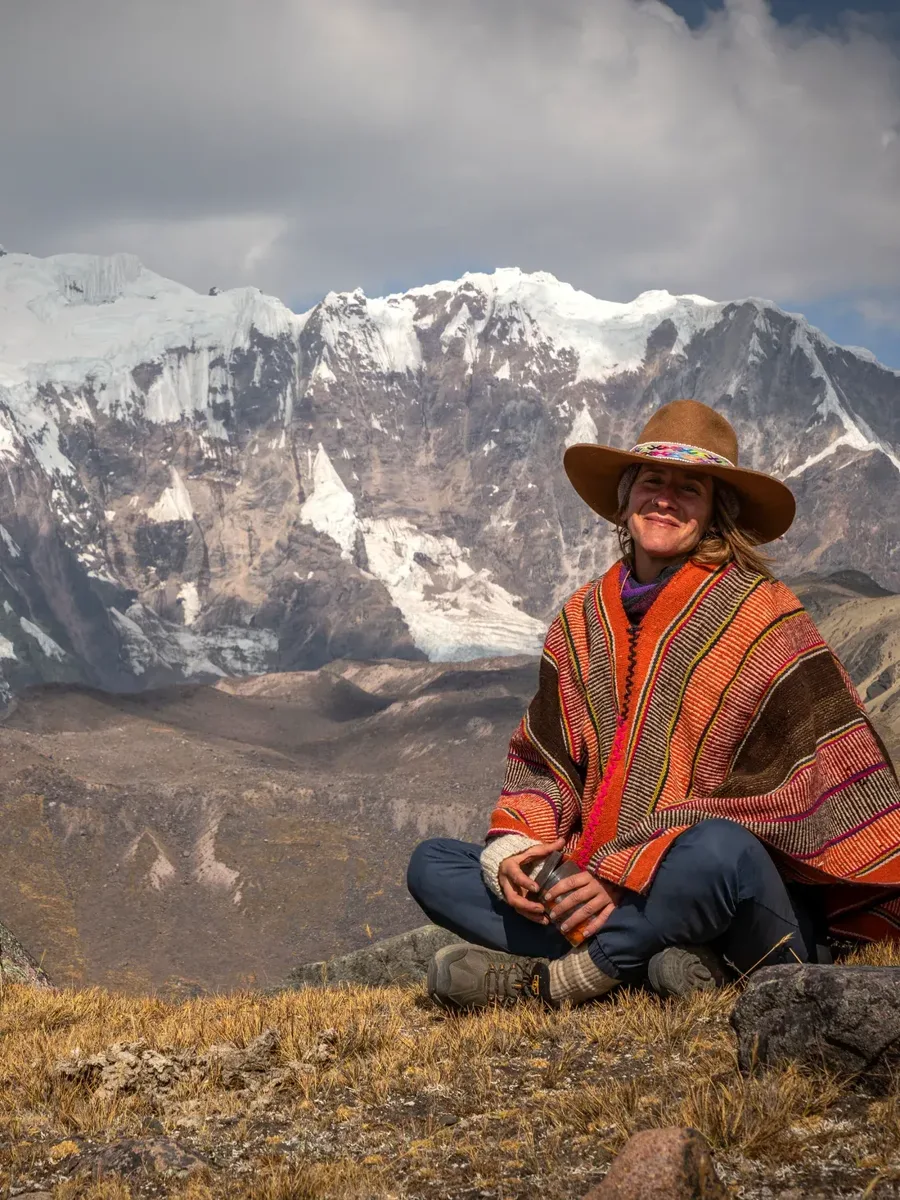 A woman in a red woven poncho and wide-brimmed hat sits in the grass, smiling, before a snow-covered range