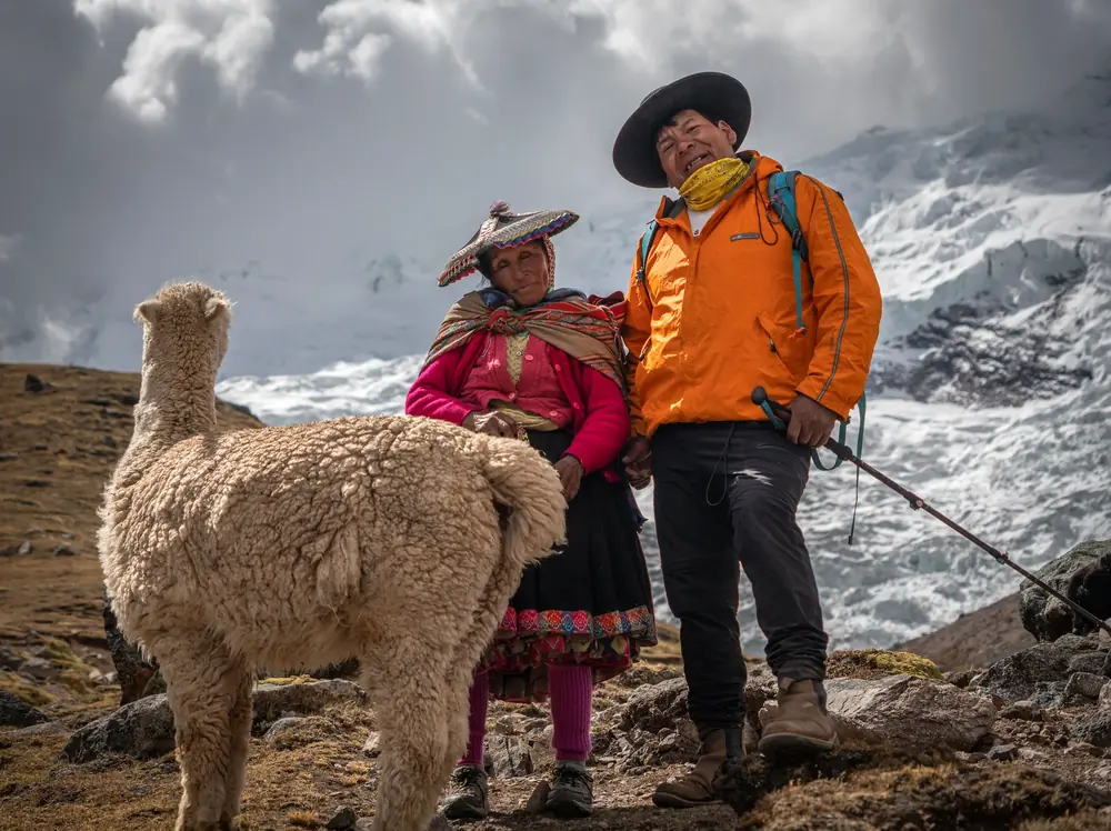 A local woman in traditional dress and a guide in an orange jacket stand beside an alpaca with a glacier behind them