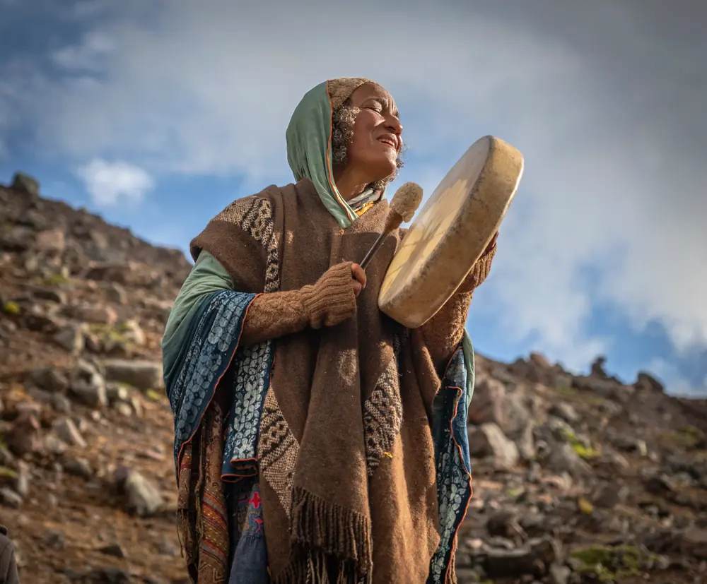 A woman in a shawl plays a frame drum under the open sky