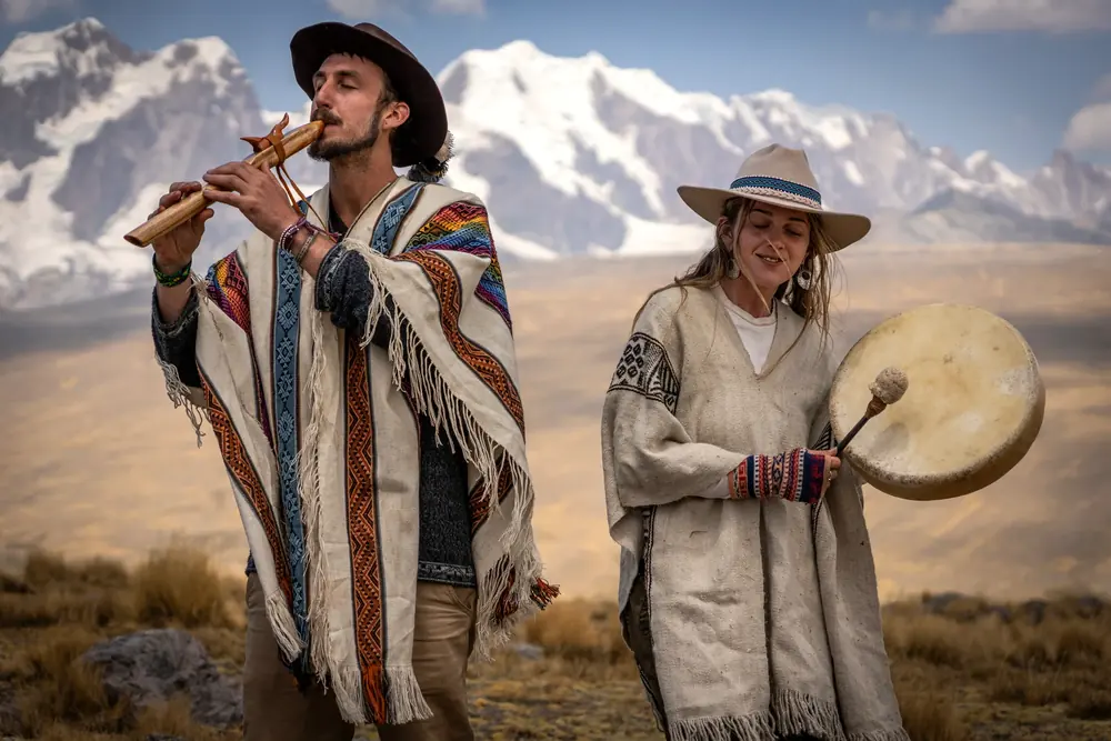 A man playing a wooden flute and a woman holding a frame drum, both in alpaca ponchos, with mountains behind
