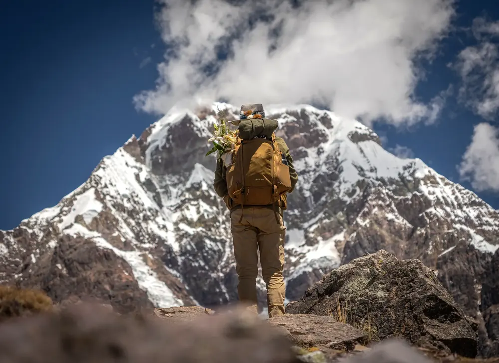 A pilgrim carrying a pack and flowers walks toward a glaciated peak