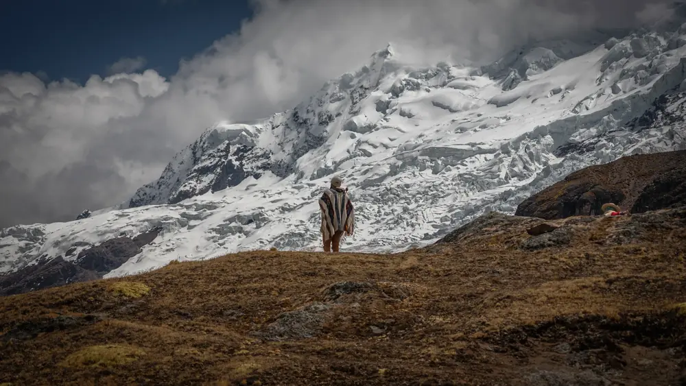 A lone pilgrim in a poncho stands on a ridge beneath a snow-covered mountain