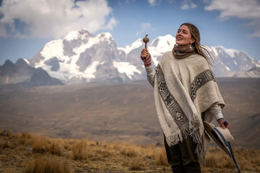 A woman in a woven poncho lifts a rattle toward the sky in front of a snowy peak