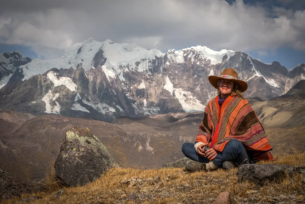A woman in a red poncho and wide-brimmed hat sits in the grass before a snowy range