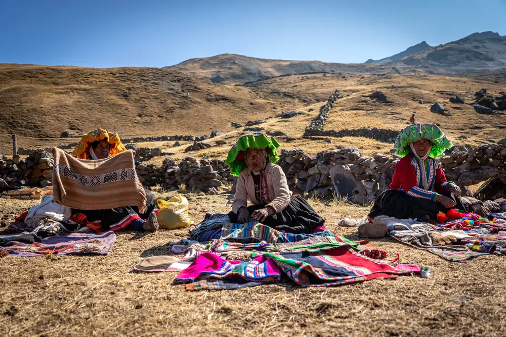 Two women in green hats sit with brightly colored woven textiles spread on the ground