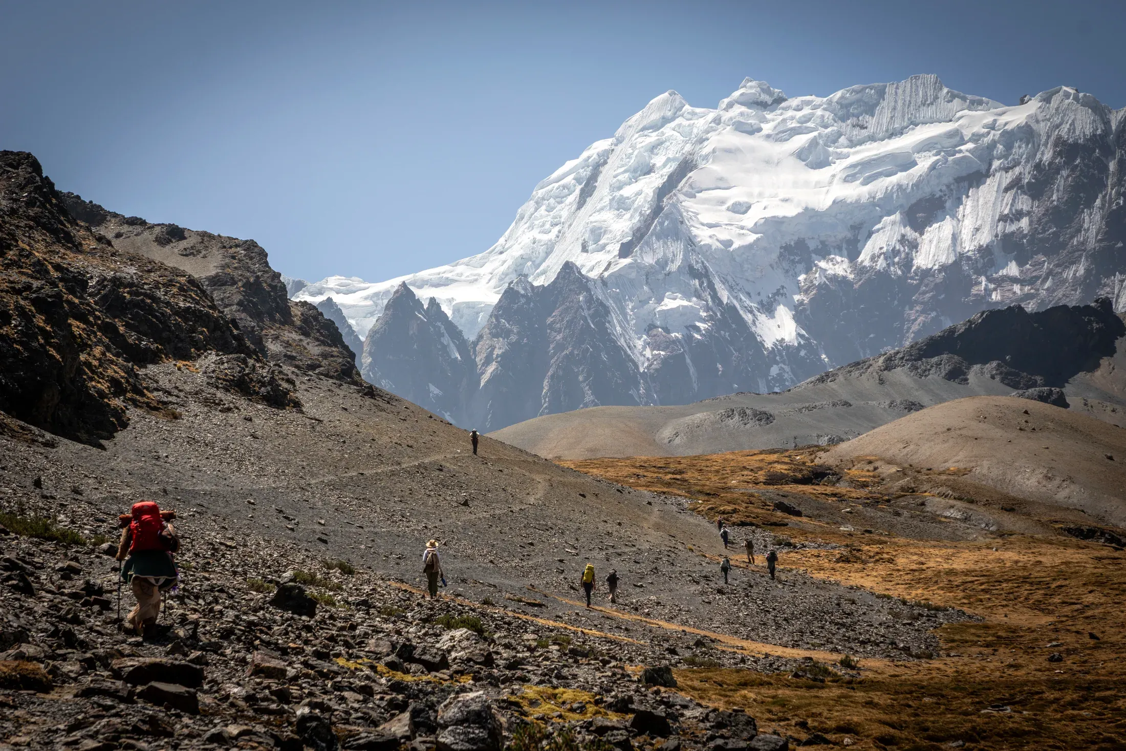 Pilgrims walking single file down a mountain trail toward a wall of snow peaks
