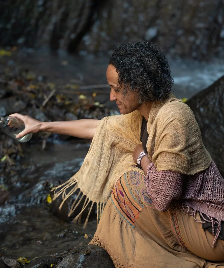 A woman kneeling on the rocks beside a stream, reaching down to the water
