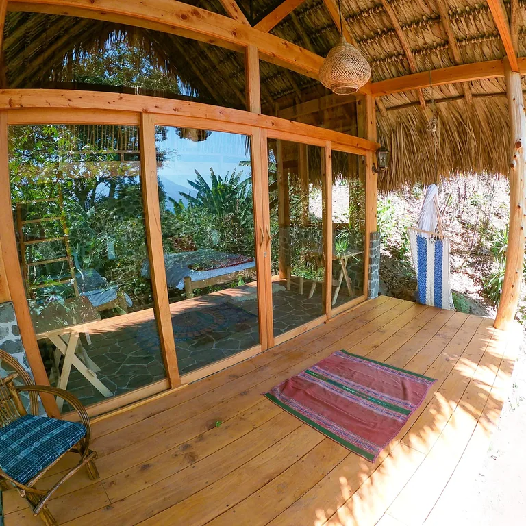 A wooden cabin porch with glass doors and a thatched roof, garden and lake view behind