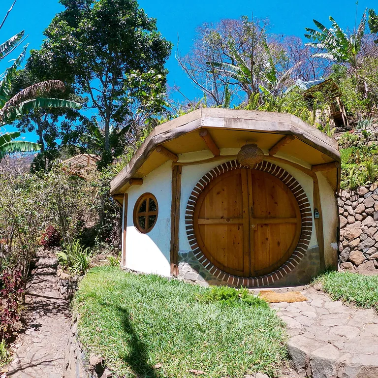 A small round cob house with a round wooden door, set in a sunny garden