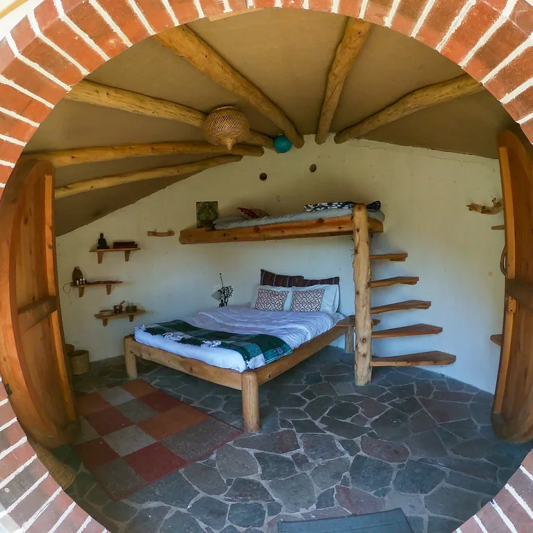 Inside a cob house at Casa Kula: a wooden bed under a curved ceiling of wooden beams, seen through the round doorway