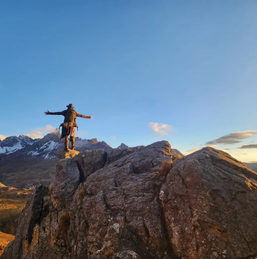 A pilgrim stands with arms outstretched on a rocky outcrop at sunset, with distant snow peaks