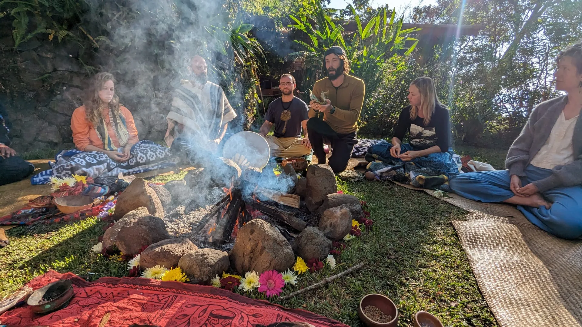 A circle of people sitting on mats around a smoking fire decorated with flowers